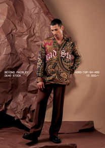 Man wearing HORD Dead Stock paisley print shirt with white gothic lettering, styled with brown trousers